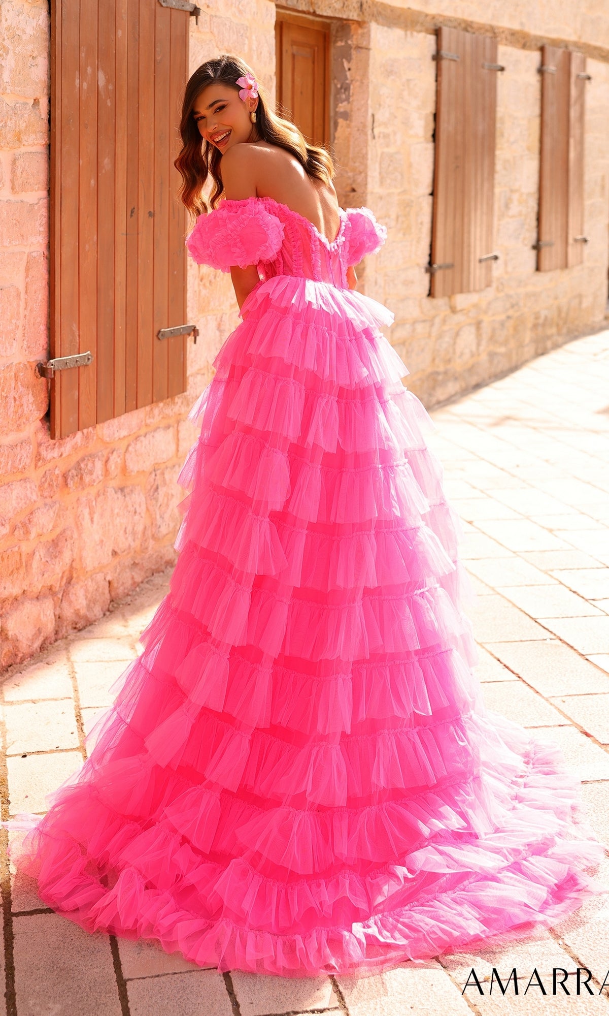 A woman in the Amarra Long Prom Dress 94000, featuring bright pink ruffled tulle and off-the-shoulder puff sleeves, stands smiling outdoors on a stone walkway, looking back over her shoulder.
